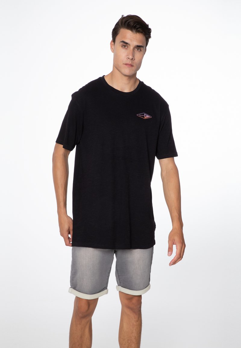 Zwarte t-shirt met korte mouwen en een klein, driehoekig logo op de linkerborst. Gecombineerd met lichtgrijze denim shorts met opgestikte zoom.