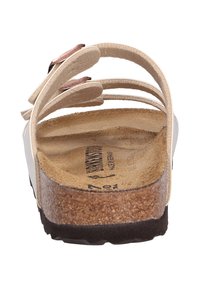 Vit sandal med flera böjda remmar, en korkfotbädd och en texturerad brun sula. Utrustad med en metallisk accent på remmen.