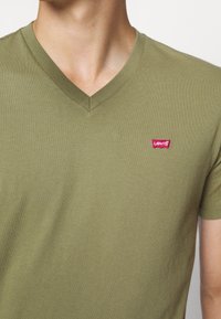 Homme portant un t-shirt Levi's col en V vert olive avec un petit logo rouge sur la poitrine gauche, visible du cou à la moitié du torse.