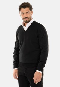 Maglione nero a scollo a V realizzato in morbido tessuto a maglia, con maniche lunghe e design aderente. Indossato sopra una camicia bianca con colletto.