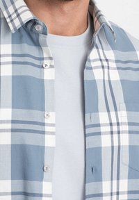 Camisa de cuadros azul claro con botones que presenta rayas blancas y azul oscuro, con un bolsillo de parche y una camiseta interior beige. Textura de tela suave.