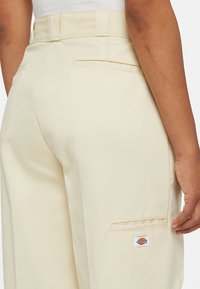 Beige katoenen broek met een rechte snit, achterzak en een klein logo op de zijkant. De stof heeft een gladde textuur.