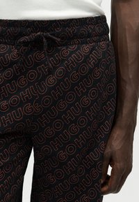 Gros plan sur un pantalon noir avec un motif répétitif du logo rouge "HUGO" et une taille à cordon, porté par une personne à la peau foncée et un haut blanc.