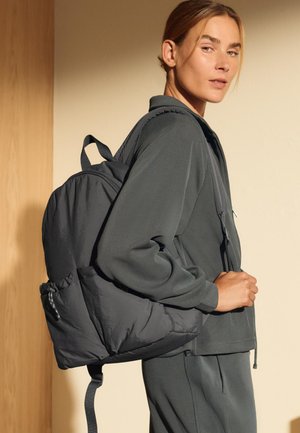Dunkelgrauer Puffer-Rucksack mit glatter Textur, gepolstertem Design, verstellbaren Trägern und äußerer Tasche. Das Modell trägt ein passendes Outfit.