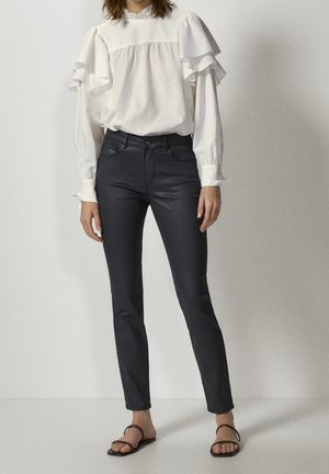 Femme portant un chemisier blanc à manches longues avec des volants et un pantalon noir slim taille haute, debout devant un fond uni.