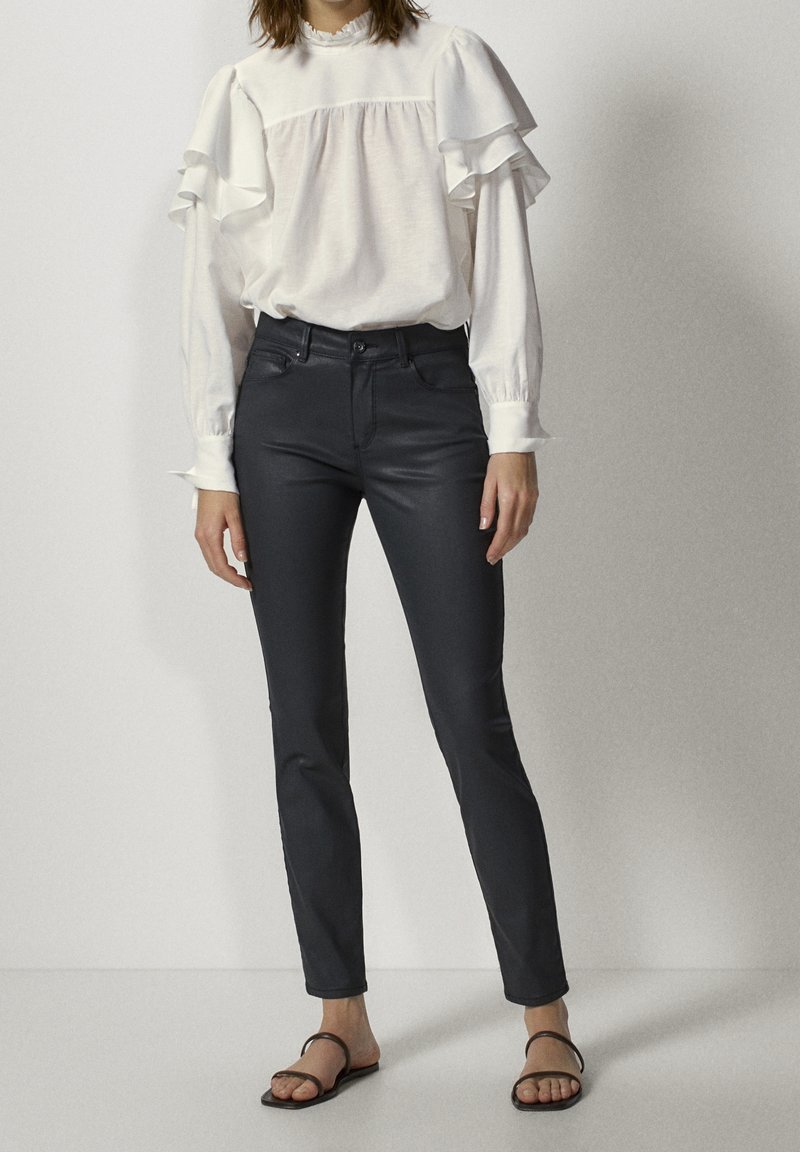 Femme portant un chemisier blanc à manches longues avec des volants et un pantalon noir slim taille haute, debout devant un fond uni.