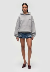 Felpa grigia oversize con tasca frontale e logo, abbinata a una mini gonna di jeans. La modella indossa tacchi a punta bordeaux. Texture liscia.