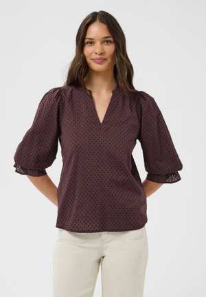 Femme aux longs cheveux bruns portant un chemisier texturé violet foncé avec des manches bouffantes et un pantalon beige clair, souriante, mains derrière le dos.