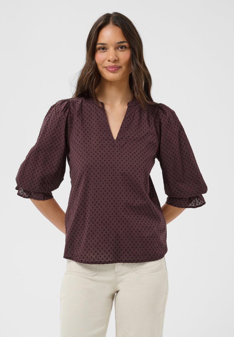 Femme aux longs cheveux bruns portant un chemisier texturé violet foncé avec des manches bouffantes et un pantalon beige clair, souriante, mains derrière le dos.