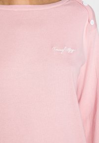 Różowy sweter z okrągłym dekoltem, prążkowaną fakturą i białym haftowanym logo. Posiada guziki na ramionach. Miękki materiał.