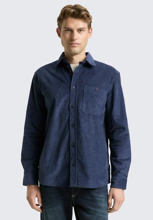 Jeune homme portant une chemise en velours côtelé bleu foncé à manches longues et à poche poitrine, se tenant devant un fond clair.