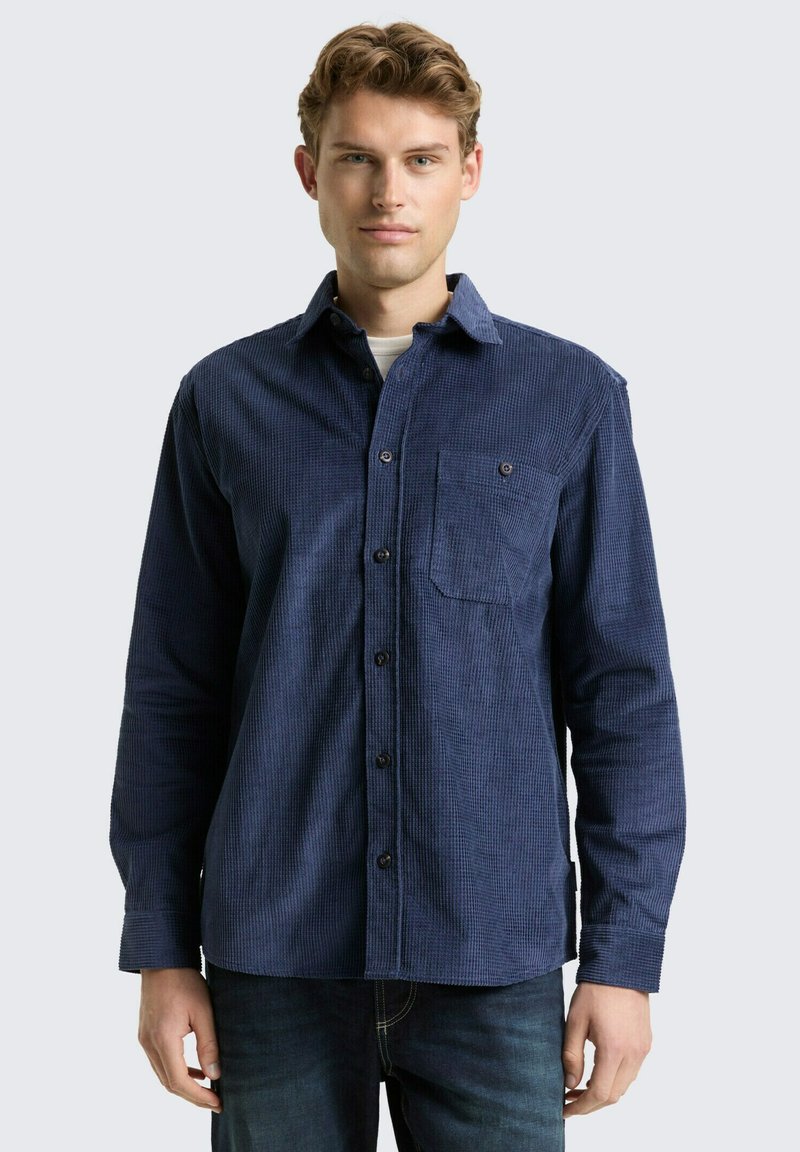 Jonge man in een donkerblauw corduroy overhemd met lange mouwen en een borstzak, staand tegen een lichte achtergrond.