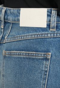 Gros plan sur la poche arrière d’un jean denim bleu avec une étiquette de marque en cuir blanc cousue à la ceinture.