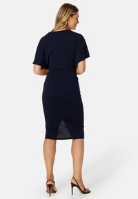 Námořnické modré šaty s krátkými volánovými rukávy, přiléhavý střih, na bocích nařasení, délka ke kolenům, decentní rozparek vzadu, doplněné černými vysokými podpatky.