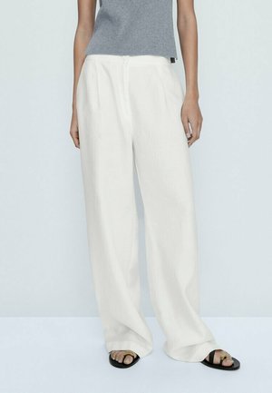 Osoba ubrana w luźne białe spodnie, szary bezrękawnik i czarne sandały, stojąca na tle jasnego, jednolitego tła.