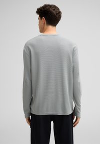 Helles graues Langarmshirt mit Waffelstruktur; runder Halsausschnitt und leicht herabgesetzte Schultern. Getragen mit dunklen Hosen.