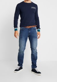 Mężczyzna noszący granatowy sweatshirt z napisem "S.O.V.R.", niebieskie dżinsy, brązowy pasek oraz czarne trampki z białymi sznurówkami na białym tle.