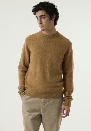 Man with short curly hair wears a tan sweater and beige trousers, standing with hands in pockets, looking to the side against a plain background.
