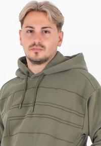 Felpa verde oliva con cappuccio e cordoncino, caratterizzata da linee cucite orizzontali sulla parte anteriore. Il materiale sembra morbido con una finitura texture.