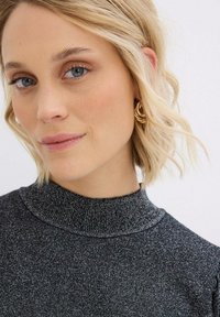 Top nero sparkly con collo alto in tessuto testurizzato. Indossato con orecchini a cerchio a spirale dorati. Capelli biondi corti e mossi.