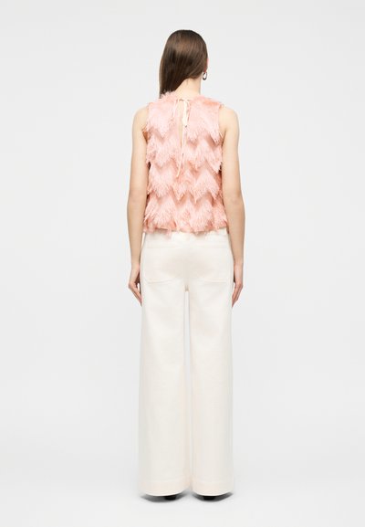 Γυναίκα με μακριά καστανά μαλλιά, ντυμένη με ροζ αμάνικη μπλούζα με υφή και κορδόνι στο πίσω μέρος και φαρδιά εκρού παντελόνια, όρθια και με πλάτη προς τα εμπρός.