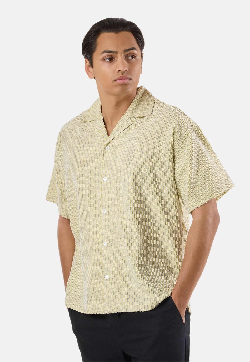 Jeune homme portant une chemise boutonnée jaune clair texturée à manches courtes, coupe décontractée, avec un pantalon noir, les mains dans les poches, regardant vers la gauche.