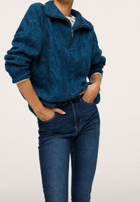 Suéter verde azulado de punto con medio cierre en el cuello, tejido texturizado y hombros caídos, combinado con jeans ajustados azules oscuros.