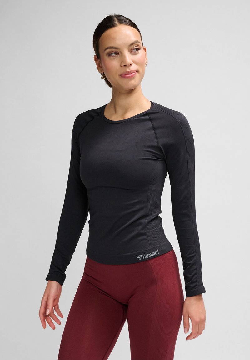 Vrouw draagt een zwart nauwsluitend topje met lange mouwen en bordeauxrode legging, staat met ontspannen handen, kijkt iets naar links.