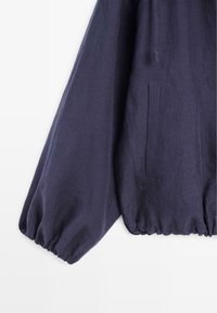 Rękaw i bok ciemnoniebieskiej kurtki z elastycznymi mankietami i pionową kieszenią na białym tle.