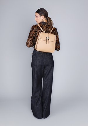 Sac à dos en cuir beige au design structuré et à fermeture à clip, porté sur le dos, associé à un haut en imprimé léopard et à un pantalon sombre ample.