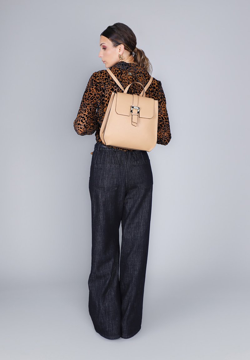 Mochila de cuero beige con un diseño estructurado y cierre de hebilla, llevada a la espalda, combinada con una blusa de estampado de leopardo y pantalones oscuros holgados.