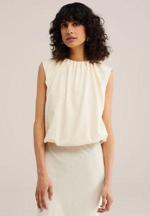 Kobieta o kręconych ciemnych włosach, ubrana w bezrękawową kremową bluzkę z marszczonym dekoltem i pasującą spódnicę, stojąca na jednolitym tle.