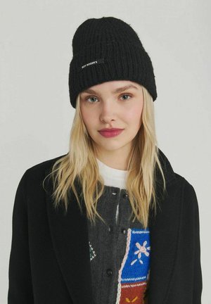 Giovane donna con lunghi capelli biondi, che indossa un berretto nero di maglia, un cappotto nero e un maglione multicolore con motivi floreali.