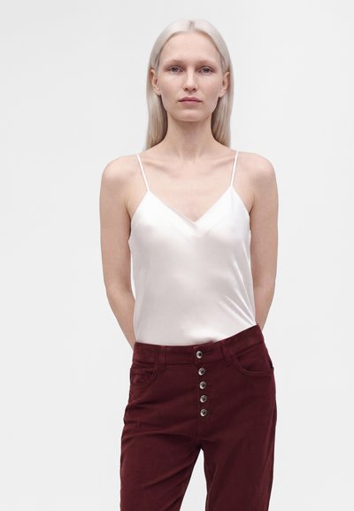 Femme blonde aux cheveux raides porte une camisole en soie blanche et un pantalon bordeaux taille haute avec cinq boutons à l'avant, debout devant un fond uni.