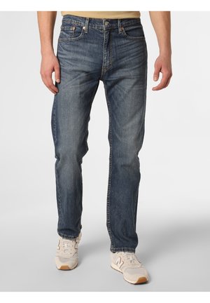 Man wearing blue jeans and white sneakers, standing with hands relaxed by his sides against a plain background.