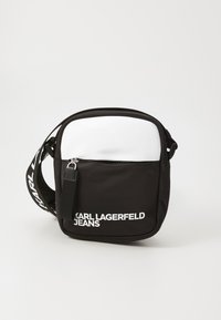 Svart og hvit budveske laget av nylon, med en glidelåsrom og fremtredende hvit logo tekst på forsiden. Justerbar stropp.
