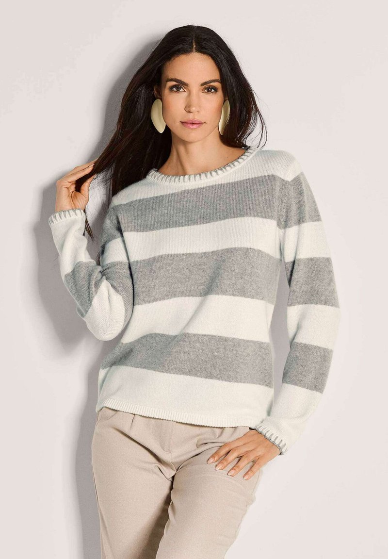 Frau mit langen, dunklen Haaren, die große cremefarbene Ohrringe und einen weiß-grau gestreiften Pullover trägt. Sie posiert mit einer Hand im Haar und der anderen auf der Hüfte.