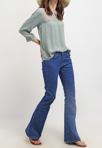 Maglietta verde chiaro a maniche lunghe con tessuto testurizzato e scollatura arrotondata, abbinata a jeans in denim blu a zampa di elefante e sandali marroni.