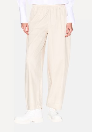 Pantaloni relaxed-fit beige chiaro con vita elastica, texture liscia e tasche laterali, abbinati a stivali bianchi con lacci spessi.
