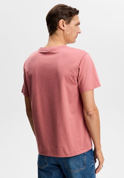 Homme portant un t-shirt à manches courtes rose poussiéreux uni et un jean bleu, vu de dos sur un fond gris clair uni.
