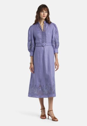 Ženska, oblečena v srednje dolgo lila obleko z nabranimi rokavi in vezenimi izrezanimi detajli, obuta v rjave sandale z visoko peto in trakovi.