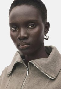 Siva jakna z zadrgo in visokim ovratnikom, izdelana iz mehke tkanine. Model nosi velike srebrne obročaste uhane. Gladka koža in nevtralen odtenek ustnic.