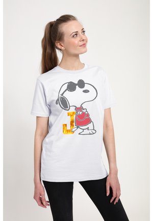 Vrouw met lang haar die een wit T-shirt draagt met Snoopy met zonnebril en een rood hemd, staand en naar boven zijwaarts kijkend.