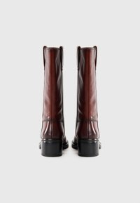 Botas de cuero marrón hasta la rodilla con un acabado suave, tacones rectangulares y costuras reforzadas a lo largo de los lados. Dos tiradores en la parte superior.