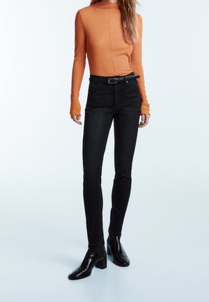 Person trägt ein enganliegendes oranges Langarmshirt, hoch taillierte schwarze Skinny-Jeans mit Gürtel und schwarze Stiefeletten, steht vor einem schlichten Hintergrund.