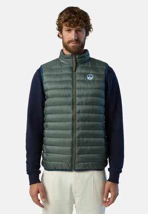 Gilet imbottito verde con colletto alto, zip frontale completa e patch con logo. Indossato sopra una maglietta a maniche lunghe blu navy. Texture liscia, design leggero.