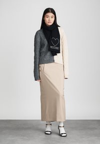 Model iført en grå og cremefarvet kabelstrikket cardigan, sort tørklæde med logo, beige nederdel med knapper og sorte hæle med hvide sokker.