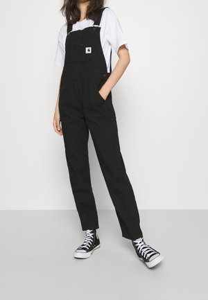 Woman wearing black overalls, white short-sleeve shirt, and black high-top sneakers standing with one hand in pocket.