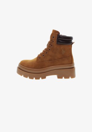 Ruskohalata ruskea nubukki nilkkabooti, jossa on paksu, teksturoitu pohja, pyöreä kärki ja pehmustettu kaulus. Varustettu metalliseoksilla ja logotiedolla sivussa.