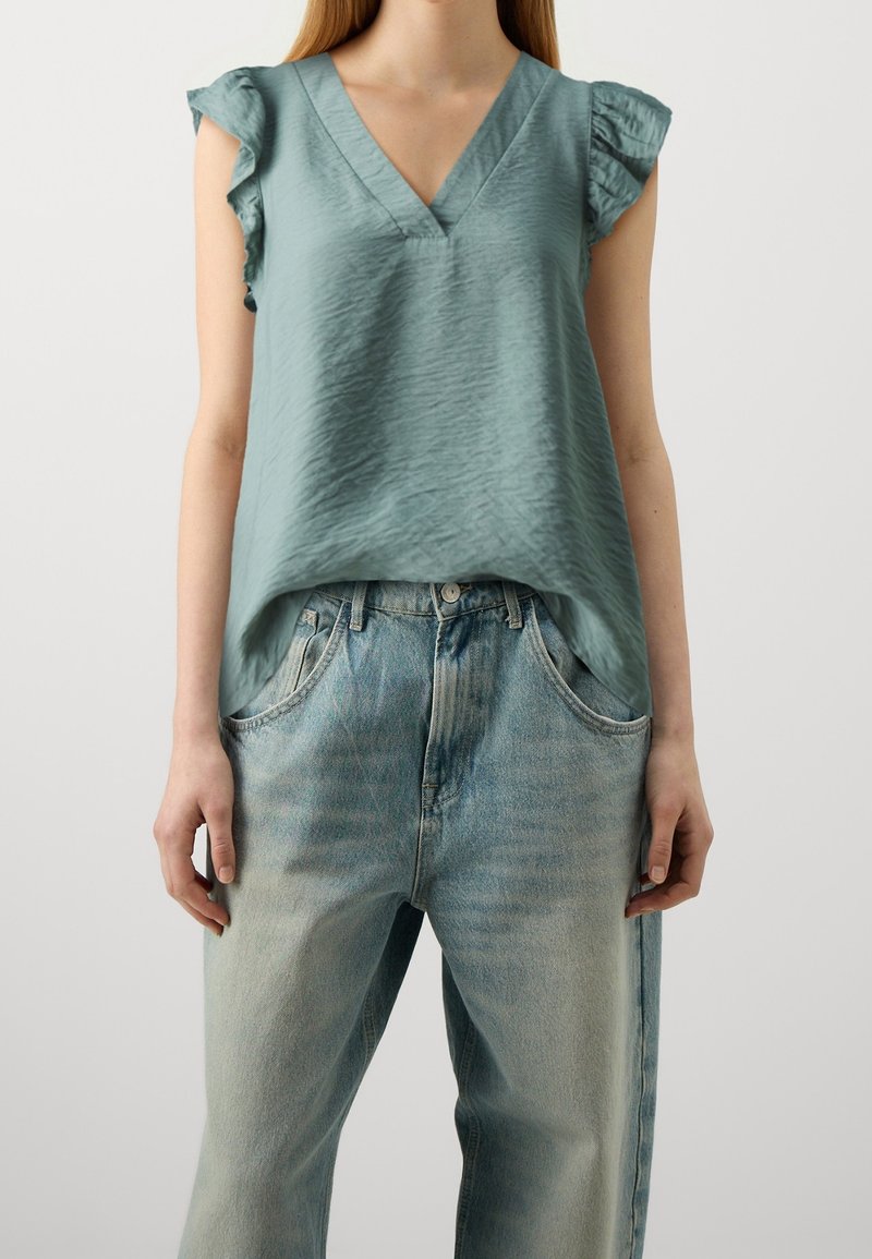 Jasnozielony bezrękawnik z dekoltem w kształcie litery V i falbaniastymi brzegami ramion, zestawiony z blednymi niebieskimi dżinsami prostymi. Teksturowana tkanina.